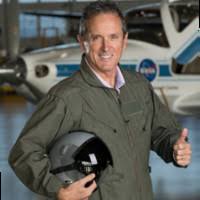Doug Trent dressed in NASA gear, holding a helmet, standing in front of a NASA aircraft, smiling and giving a thumbs up.