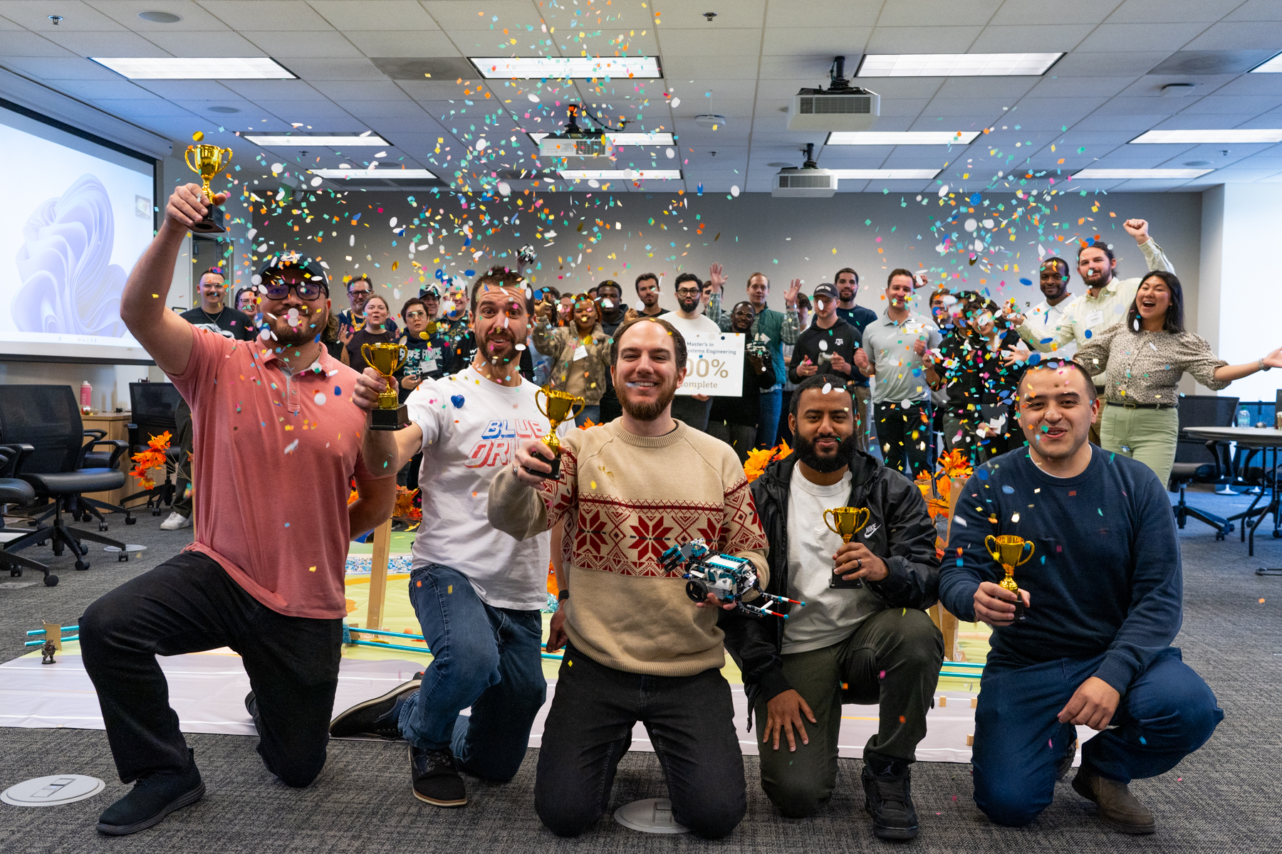 Five smiling students stand together, each holding a trophy, while the student in the center also holds a small robot.