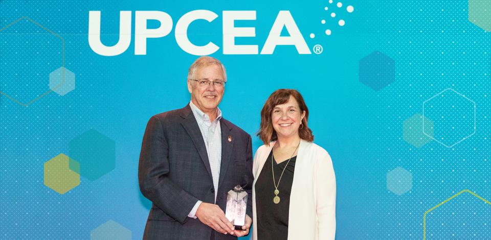 Nelson Baker in front of UPCEA branded backdrop holding award alongside the current UPCEA president.