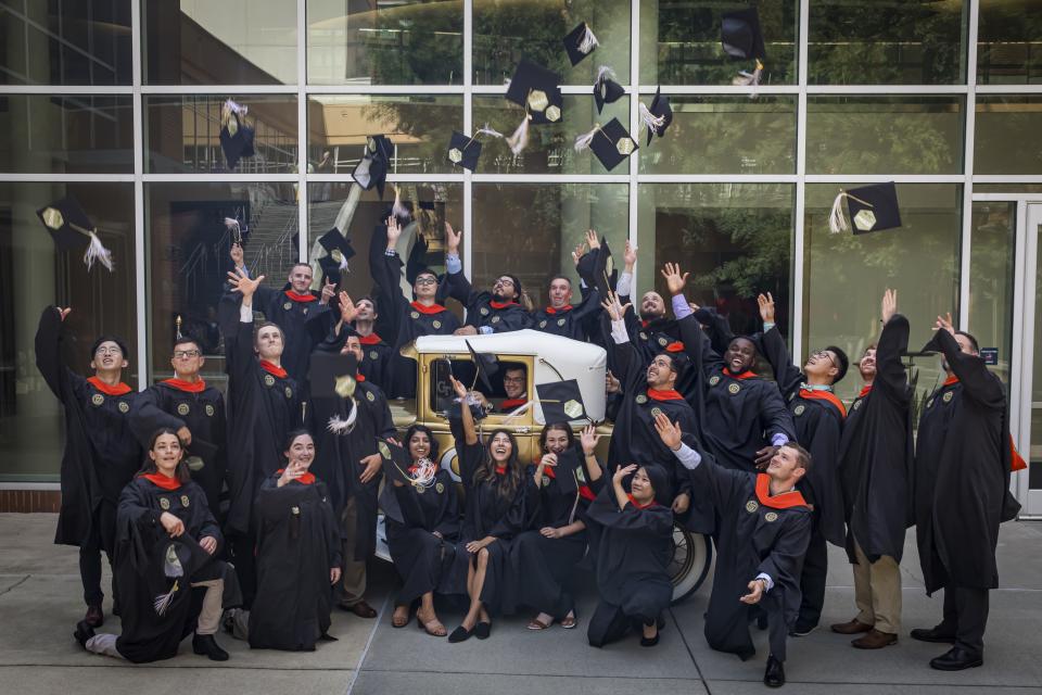 Professional Master's graduates wearing regalia standing in front of the Ramblin' Wreck tossing their caps 