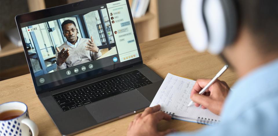 A man is taking notes while attending an online class using a laptop.