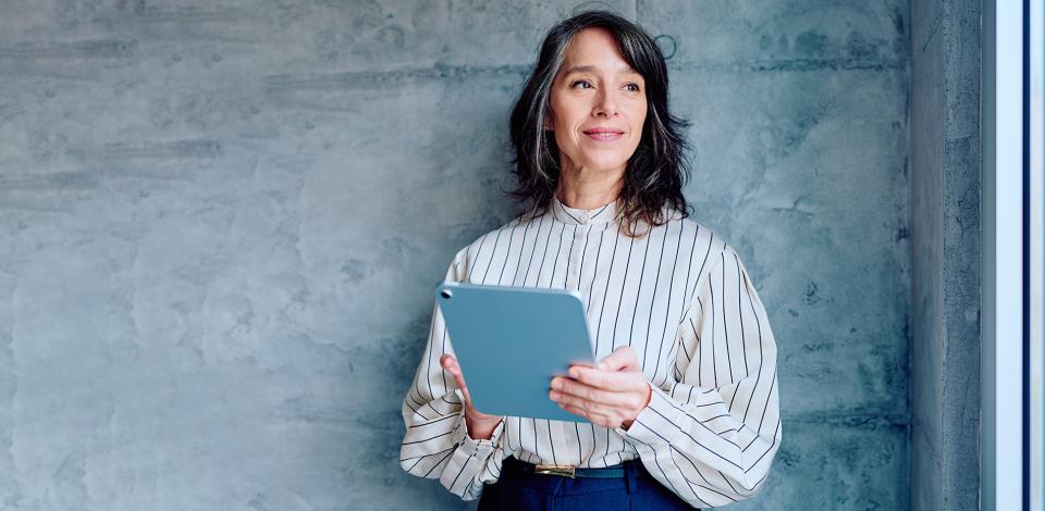 A woman holding a tablet while standing against a gray wall.