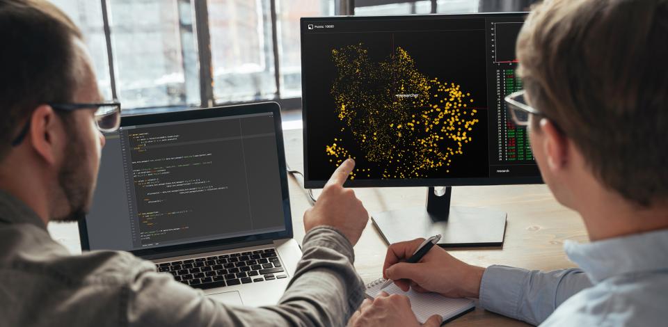 Closeup of two data science specialists looking at two computer screens, analyzing big data.
