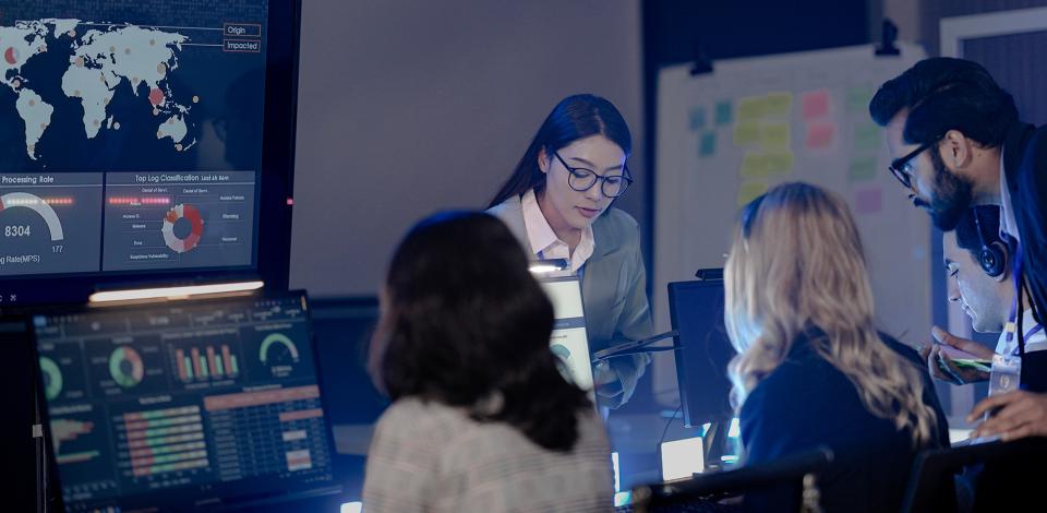 Four people working in an intelligence office viewing screens with reports and world maps