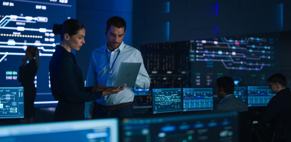 Two professionals review information on a laptop in a control room filled with screens displaying network data and system dashboards.