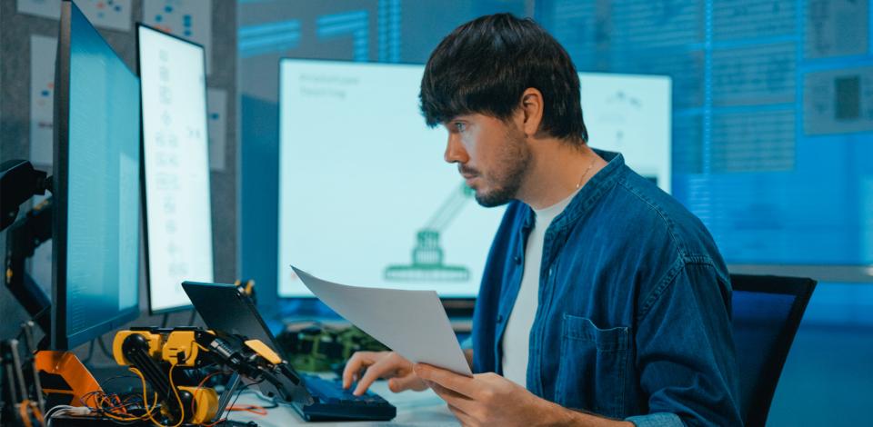 A man working at a desk with multiple computer monitors displaying data and charts, holding a document while focused on the screen.