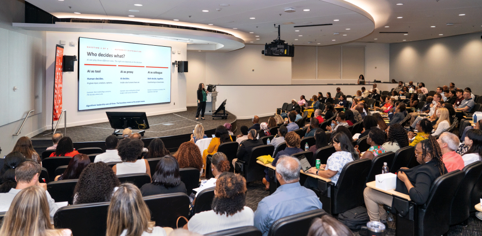 Audience members attend a professional development presentation in a large auditorium while a speaker discusses AI decision-making.