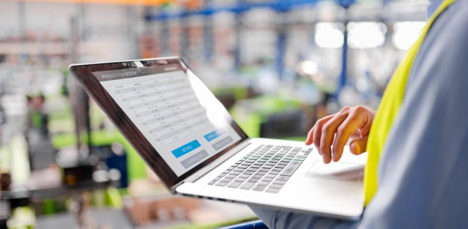 A person wearing a safety vest uses a laptop to review operational data in a manufacturing facility.