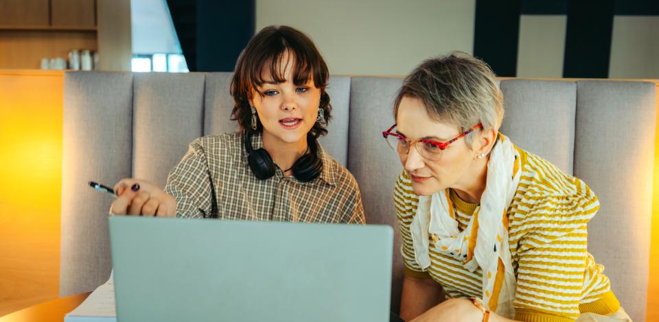 Two colleagues from different generations collaborate around an open laptop in an office.
