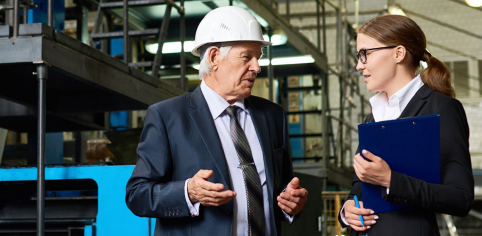 Man wearing hard hat talking to woman in front of factory equipment