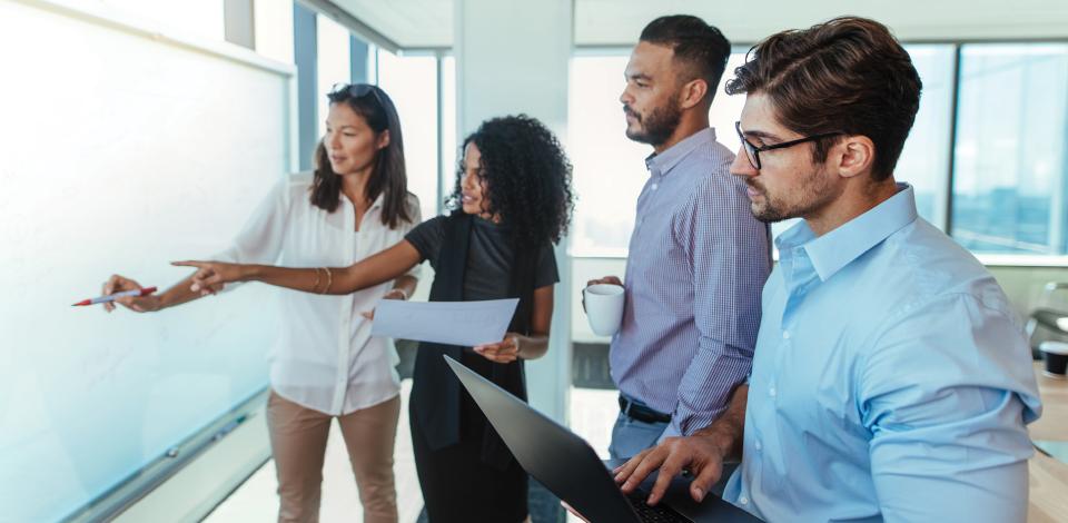 Two women and two men are collaborating in a business setting. All four are facing a whiteboard and look engaged while one woman points to the board.