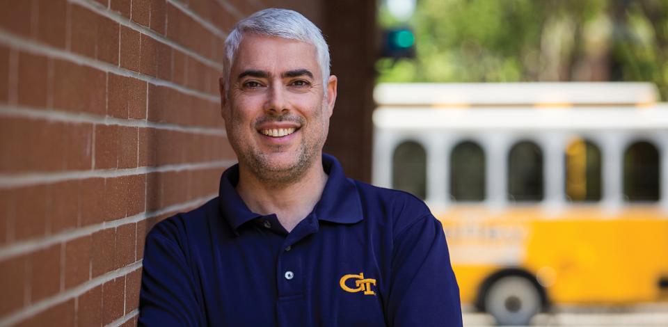 Joel Sokol smiles while he leans against a brick wall. The Georgia Tech Trolley passes in background.
