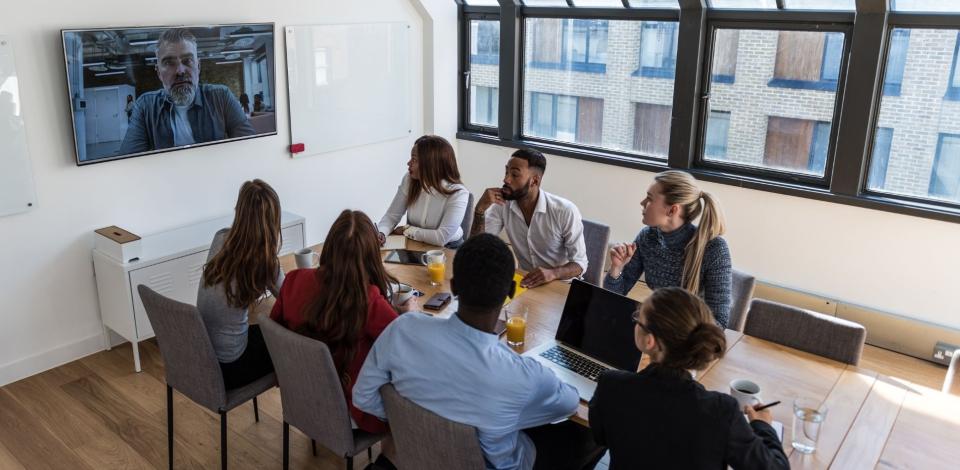 Working professionals conducting a hybrid business meeting, with some colleagues in a conference room and one colleague on video conference.