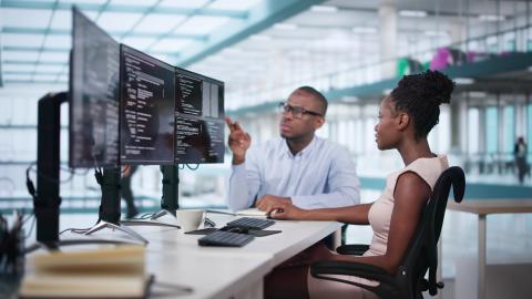 Woman sits in front of three monitors reviewing Python code with her male coworker for the FlexStack in Python AI Principles Certificate.
