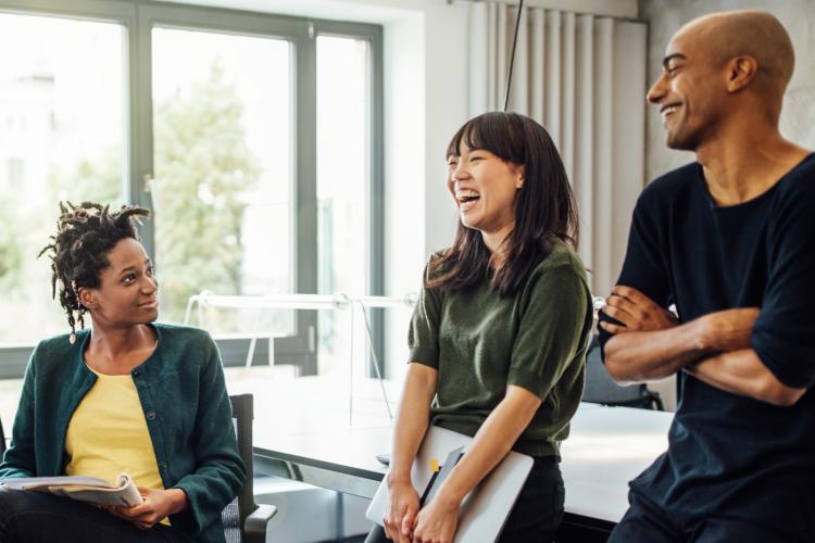 A group of colleagues gathered in a conference room or work area talking and smiling. 