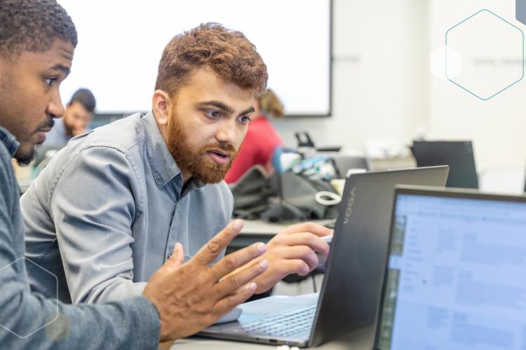 Two men sitting at a desk and looking at a laptop screen.