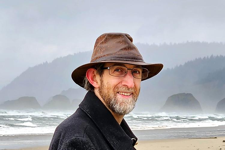 A headshot of Scott Cardinal wearing a coat and brimmed hat on a foggy beach