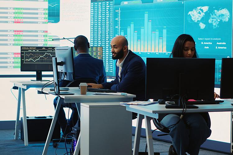 Three people in suits sit at desks in an open office space with screens displaying charts and a world map.