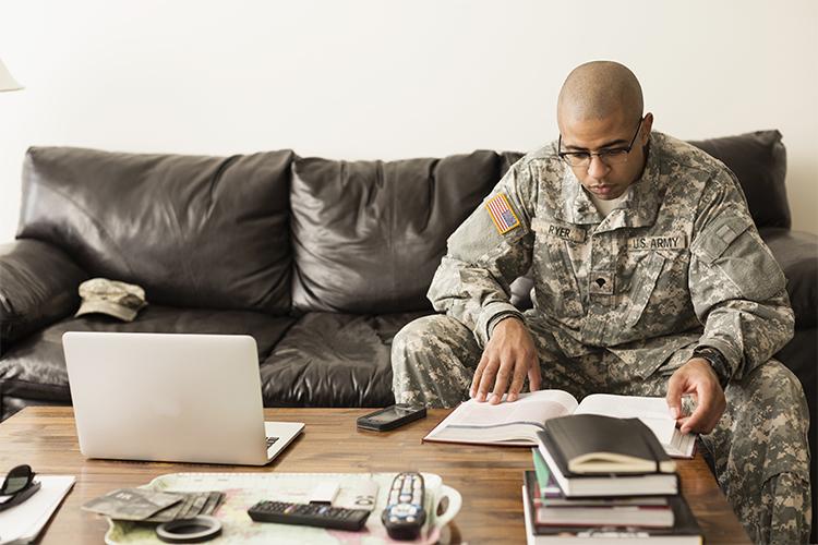Individual in military uniform reviewing materials and a laptop while sitting on a couch.