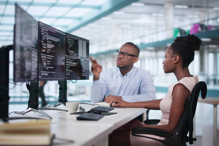 Woman sits in front of three monitors reviewing Python code with her male coworker for the FlexStack in Python AI Principles Certificate.