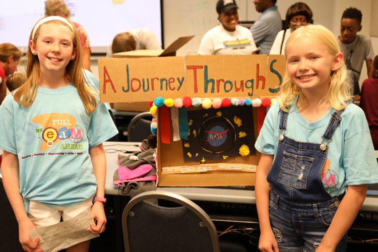 Two children standing beside a cardboard project display titled ‘A Journey Through S’ decorated with colorful pom-poms and NASA logo.
