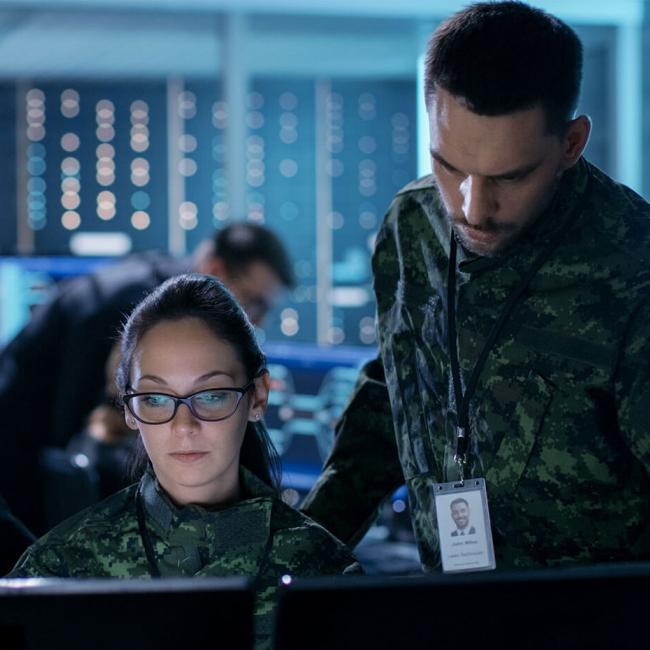 A man and woman in military fatigues accompanied by a male government official looking at a computer in a control room.