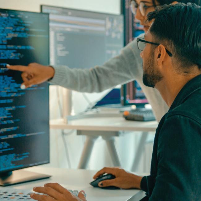 A person pointing at a computer screen and a person sitting in front of it