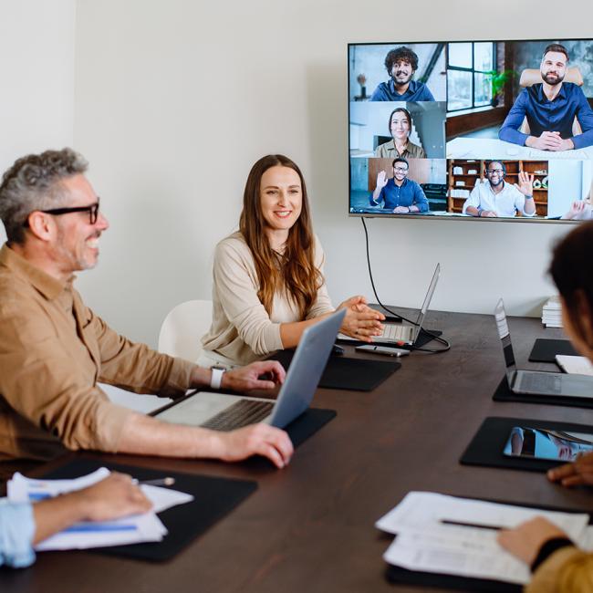 A group of coworkers are holding a meeting in a conference room while several of their colleagues are attending it virtually through a large computer screen.