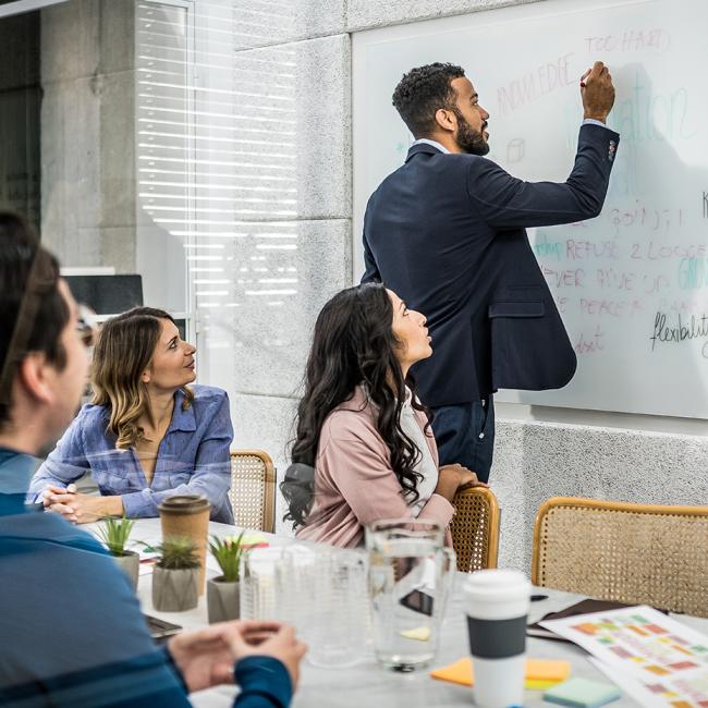 Man writing on a whiteboard with colleagues watching.