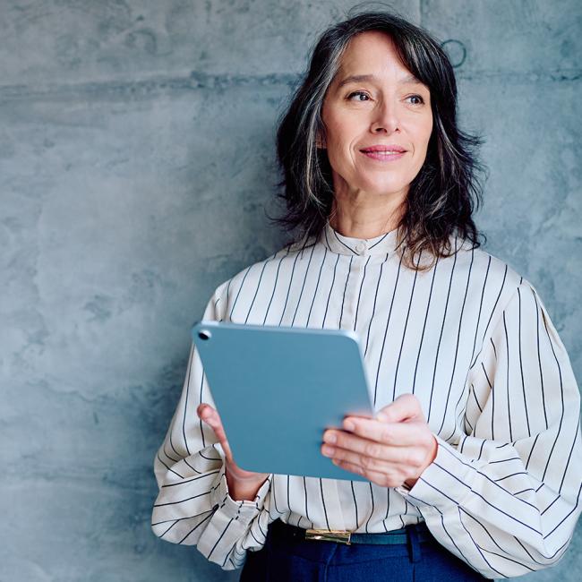 A woman holding a tablet while standing against a gray wall.