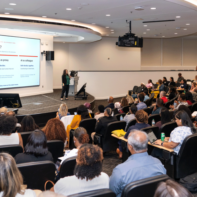 Audience members attend a professional development presentation in a large auditorium while a speaker discusses AI decision-making.