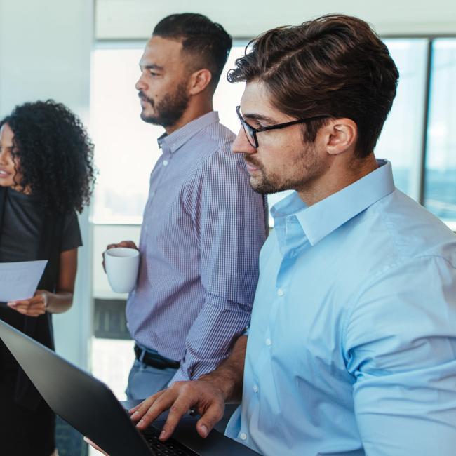 Two women and two men are collaborating in a business setting. All four are facing a whiteboard and look engaged while one woman points to the board.